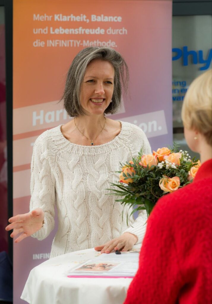 Coach Maria Depker steht an einem runden, weißen Stehtisch und spricht zu einer Frau ihr schräg gegenüber. Auf dem Tisch steht ein Strauß mit orangen Rosen. Die zweite Person ist nur von hinten zu sehen. Im Hintergrund ist ein orange-blauer Banner zu sehen.